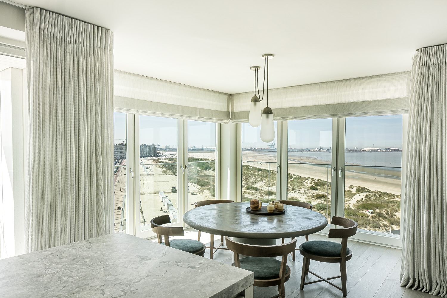 Dining area with round table, cushioned chairs, pendant light, and panoramic view of beach and coastline.