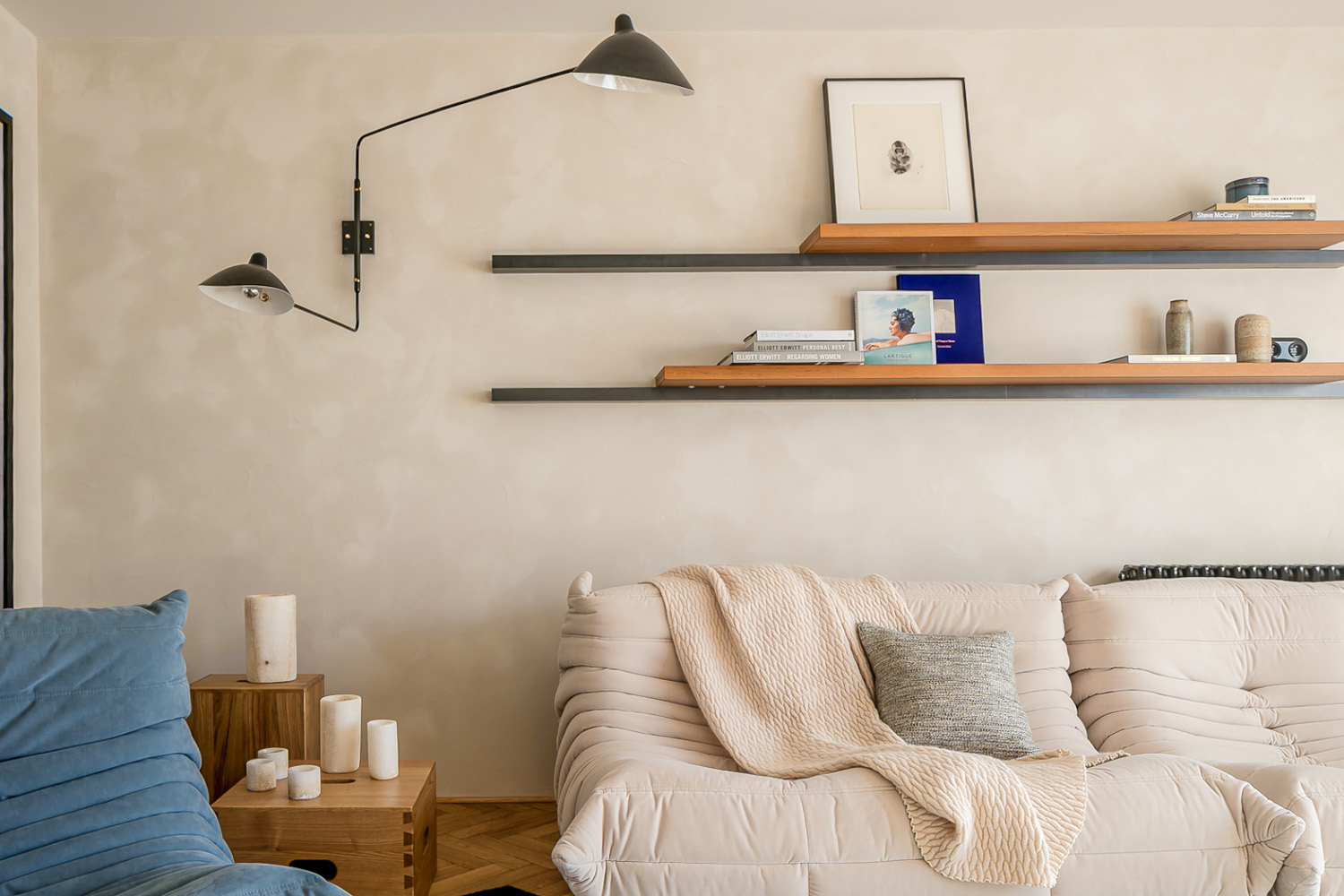 Living room with beige tufted sofa, blue chair, wooden side tables with candles, wall-mounted lamps, and floating shelves.