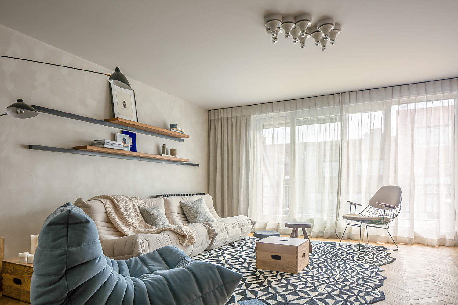 Living room with beige sofa, blue lounge chair, geometric rug, floating shelves with books and art, and wire-frame chair near window.