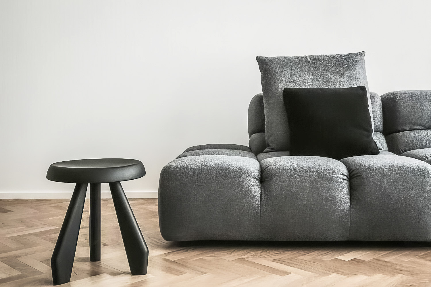 Tufted grey sofa with black pillow, black stool, and herringbone wood floor against white wall.