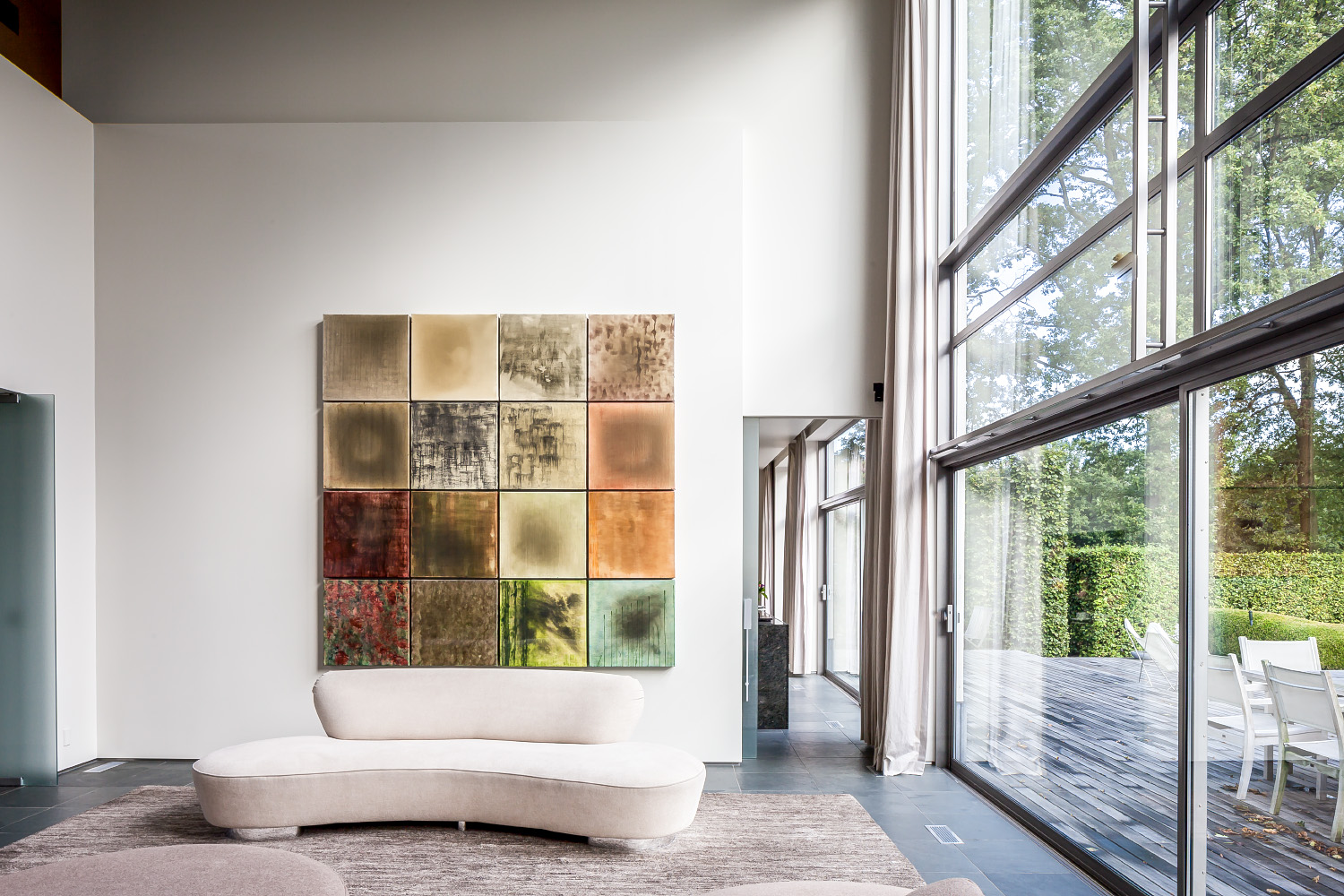 Living room with curved cream sofa, textured rug, glass wall, outdoor deck, and 20-panel art installation.
